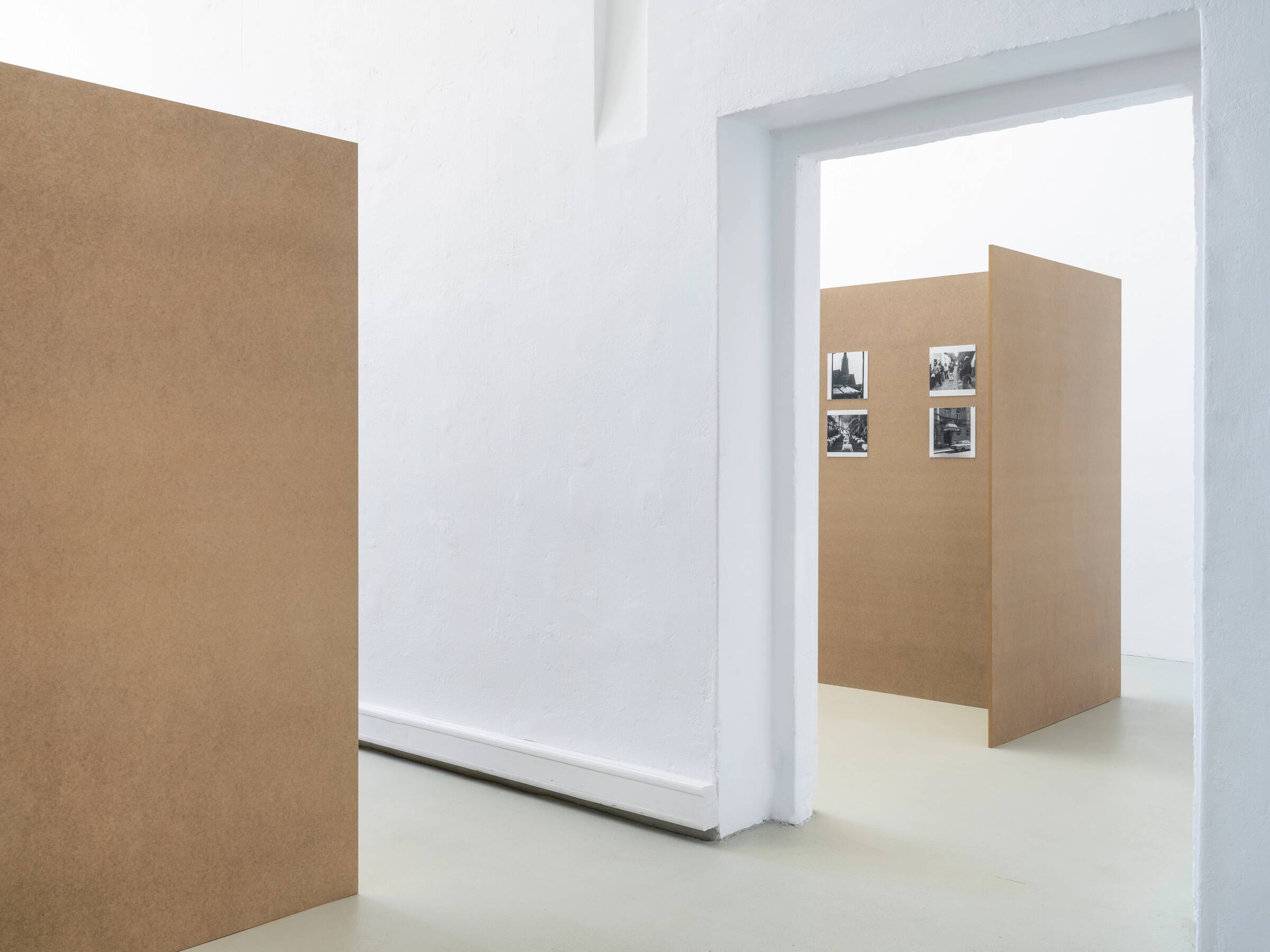 Raum mit weißen Wänden und grauem Boden, darin mehrere braune Stellwände, an einer sind vier schwarz-weiße Fotografien in zwei Reihen angebracht.
