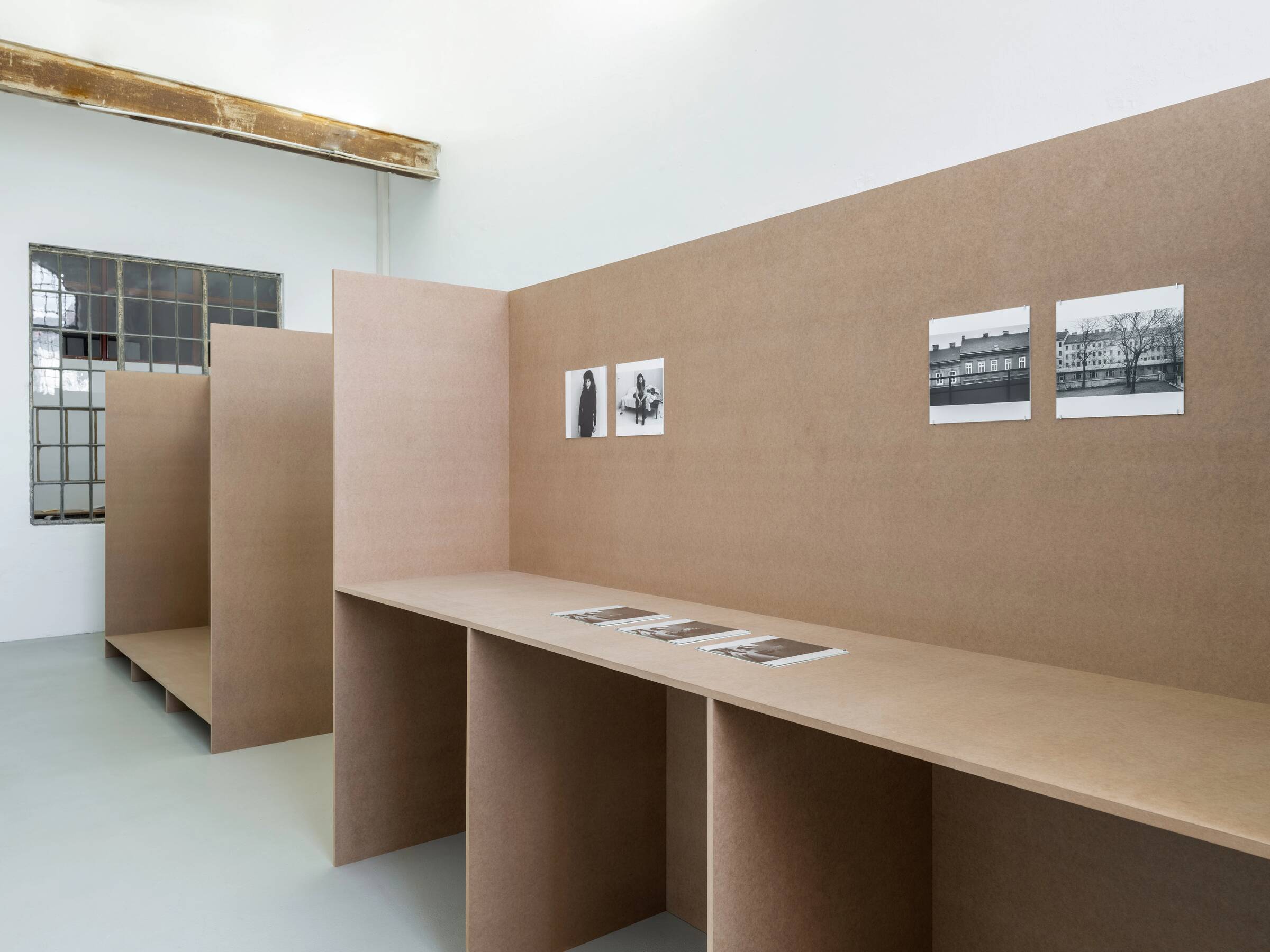 Ausstellungsansicht mit braunen Stellwänden, auf denen vier schwarz-weiße Fotografien hängen, und mehreren schwarz-weißen Fotografien auf einer Ablage.