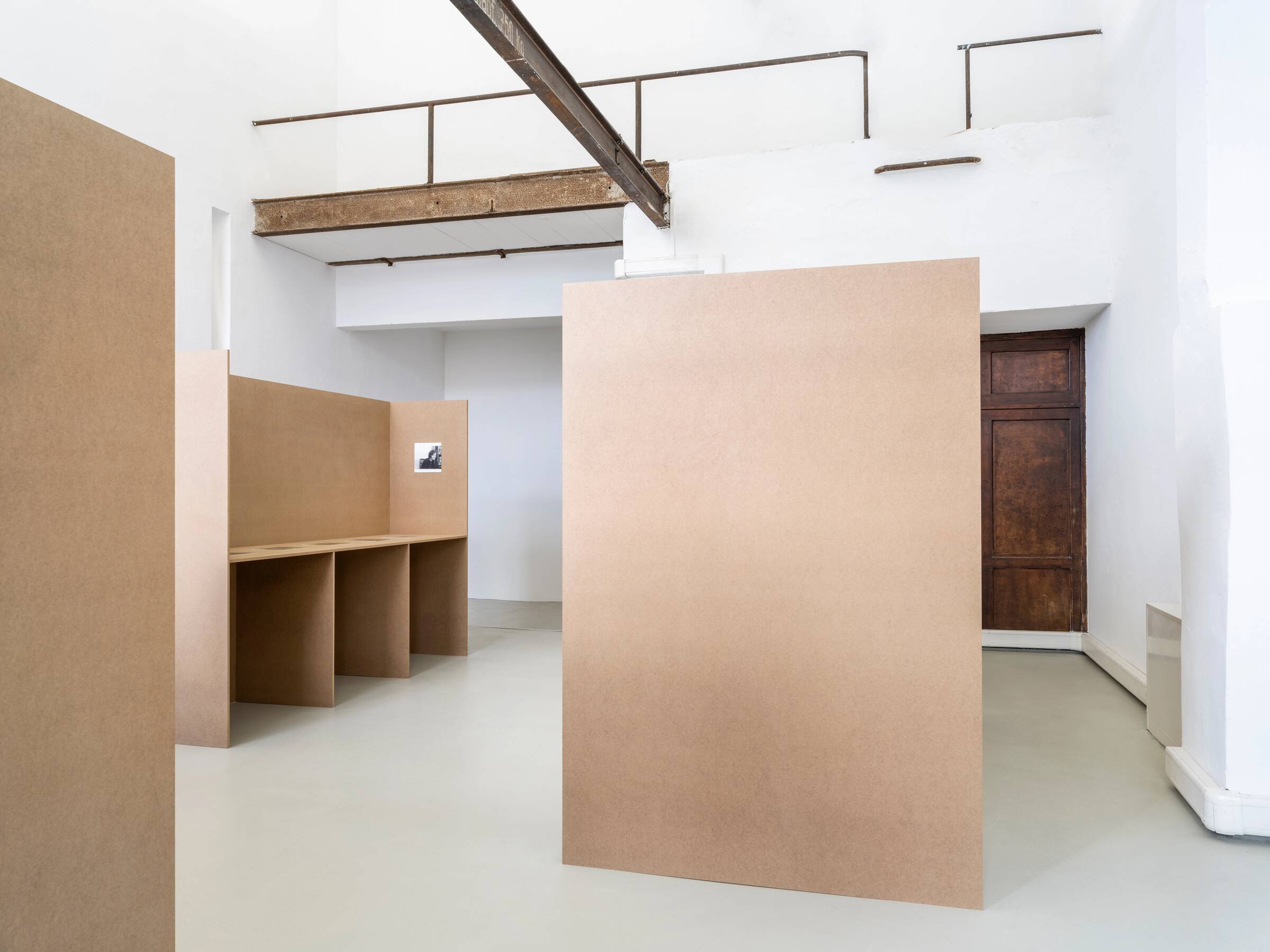 Leere Ausstellungsräume mit hohen weißen Wänden, braunen Trennwänden und einer Holztür im Hintergrund.