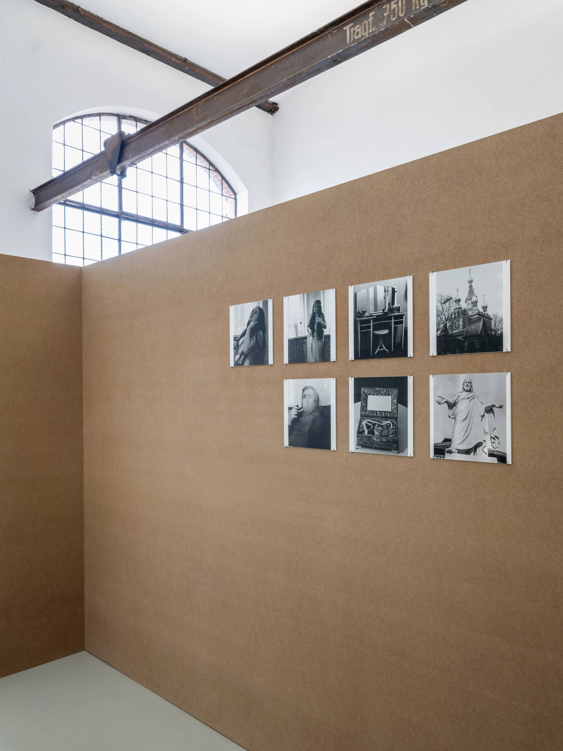 Sieben schwarz-weiße Fotografien in zwei Reihen an einer braunen Wand in einer Ausstellung.