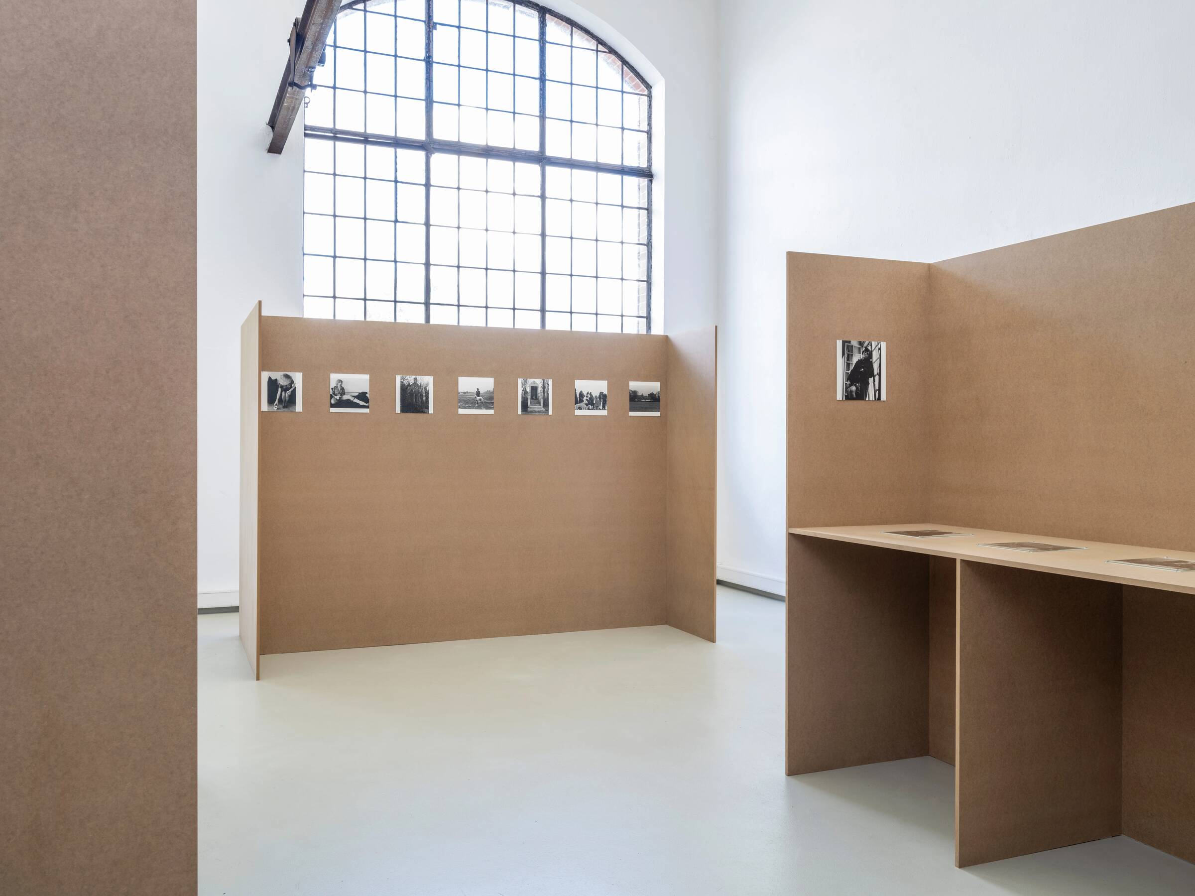 Ausstellungsansicht mit braunen Stellwänden, auf denen mehrere schwarz-weiße Fotografien hängen, und einem großen Fenster im Hintergrund.