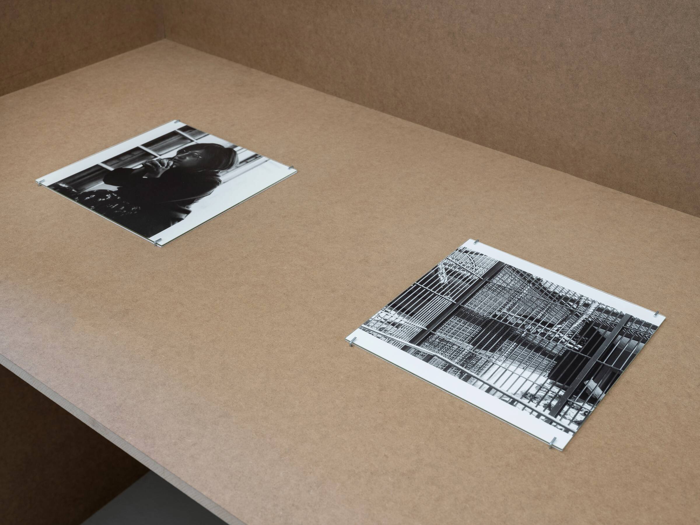 Zwei schwarz-weiße Fotografien liegen auf einer braunen Tischplatte, eine zeigt eine Person mit Kamera, die andere ein Gitter oder Zaun.