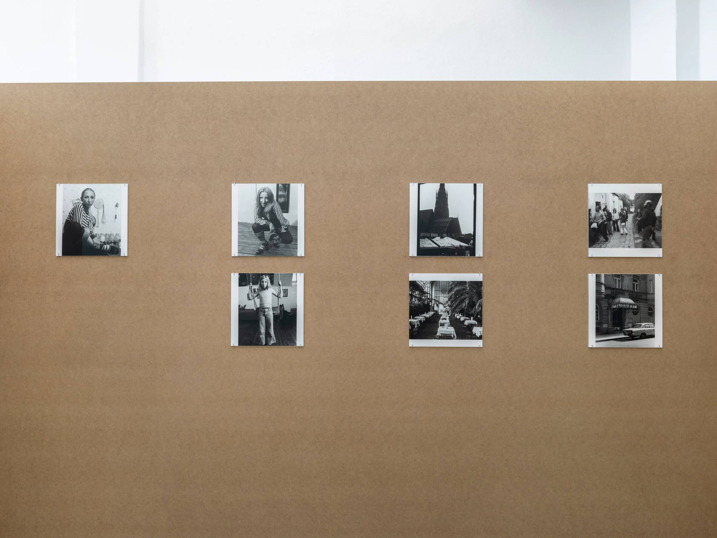 Sieben schwarz-weiße Fotografien hängen an einer braunen Wand, zeigen Porträts und Stadtansichten in einer Ausstellung.