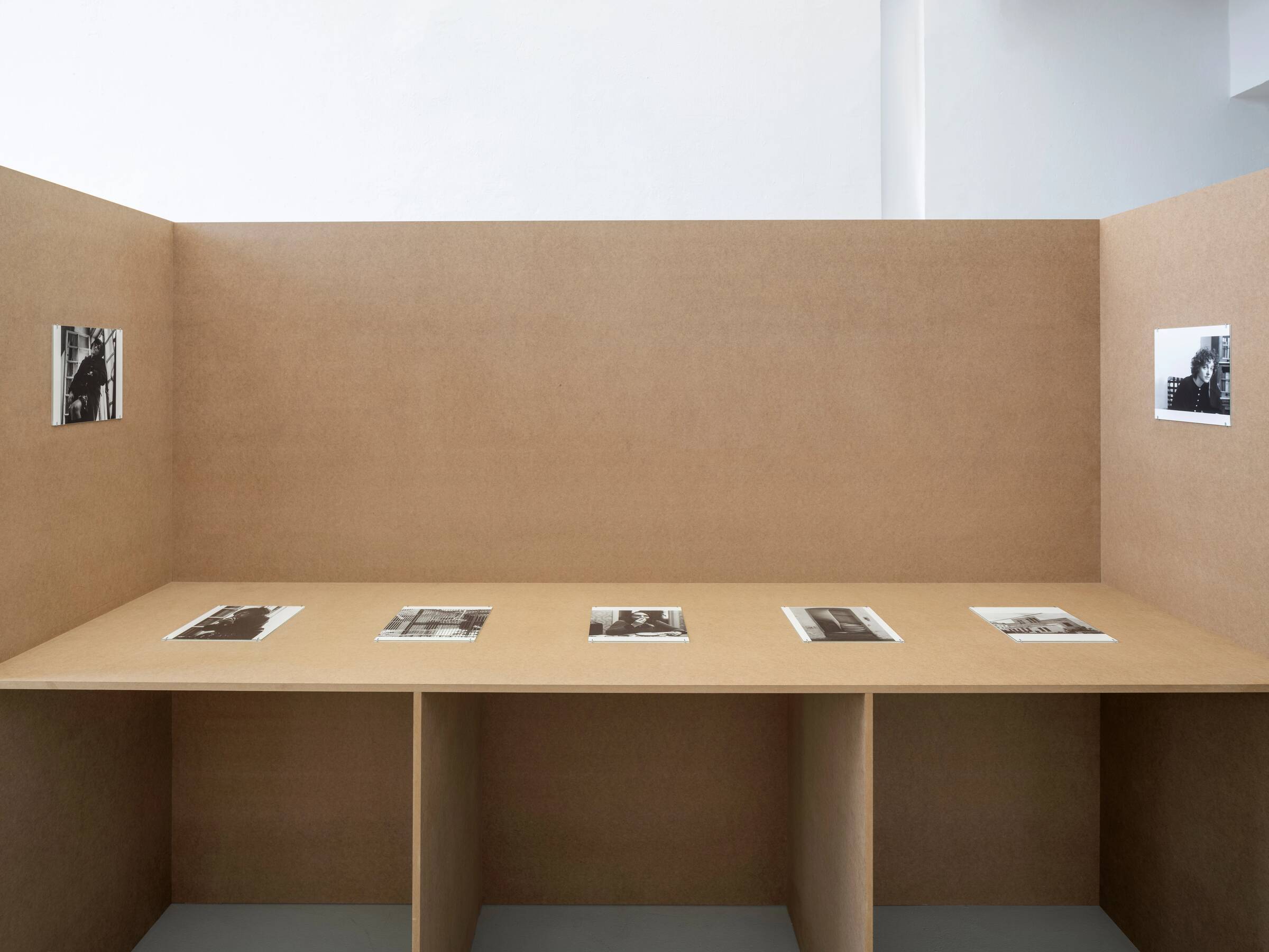 Ausstellungsansicht mit sechs schwarz-weißen Fotografien auf braunen Stellwänden, zwei hängen seitlich, vier liegen flach auf einer Ablage.
