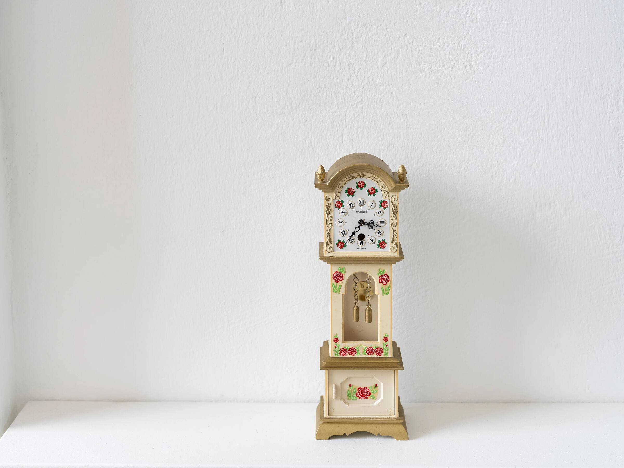 Eine kleine Standuhr aus Holz mit einem cremefarbenen Gehäuse, das mit roten Rosen und grünen Blättern bemalt ist. Das Zifferblatt ist weiß mit schwarzen arabischen Zahlen und schwarzen Zeigern. Unter dem Zifferblatt befindet sich ein kleines Fenster mit sichtbaren Pendelgewichten. Die Uhr steht auf einer weißen Oberfläche vor einer weißen Wand.