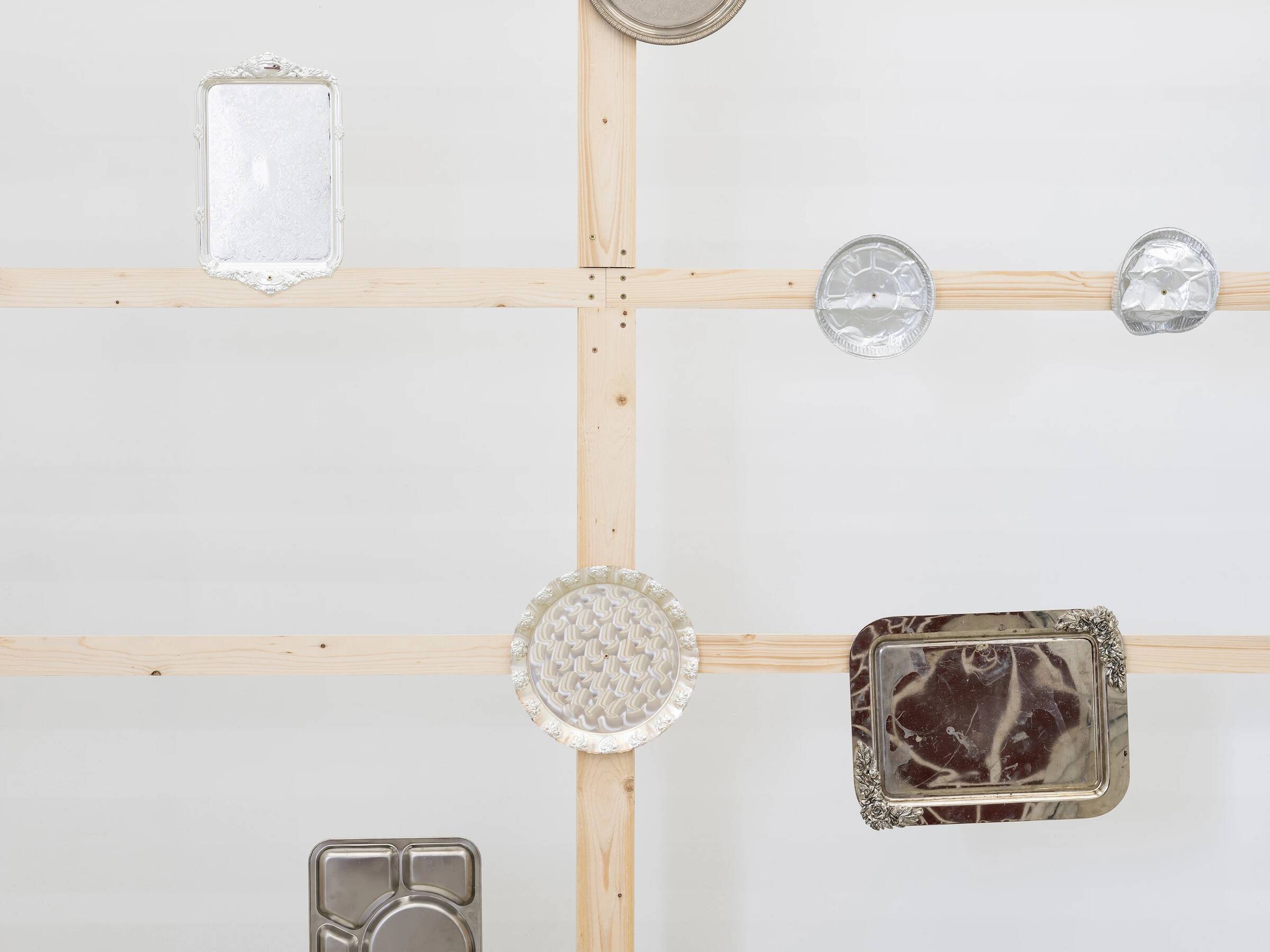 Mehrere unterschiedlich geformte und große silberne Tabletts und Schalen sind an einer weißen Wand mit sichtbaren Holzlatten befestigt.