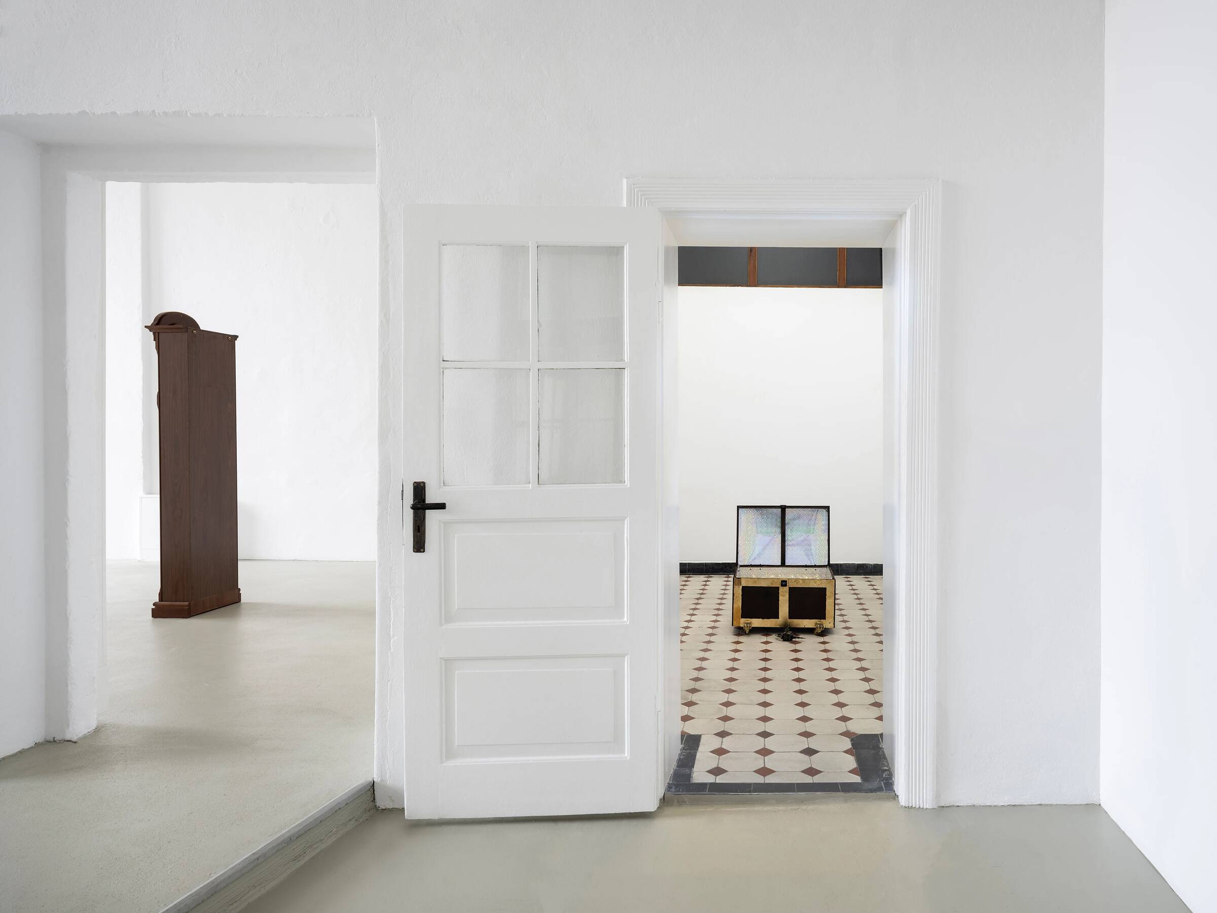Blick durch eine offene weiße Tür mit vier Fenstereinsätzen in einen Raum mit schwarz-weiß gemustertem Boden. Im Raum steht eine niedrige, rechteckige Holztruhe mit zwei Bildschirmen darauf. Links im Bild ist eine hohe, braune Standuhr in einem angrenzenden Raum zu sehen. Die Wände sind weiß, der Boden im Vordergrund hellgrau.