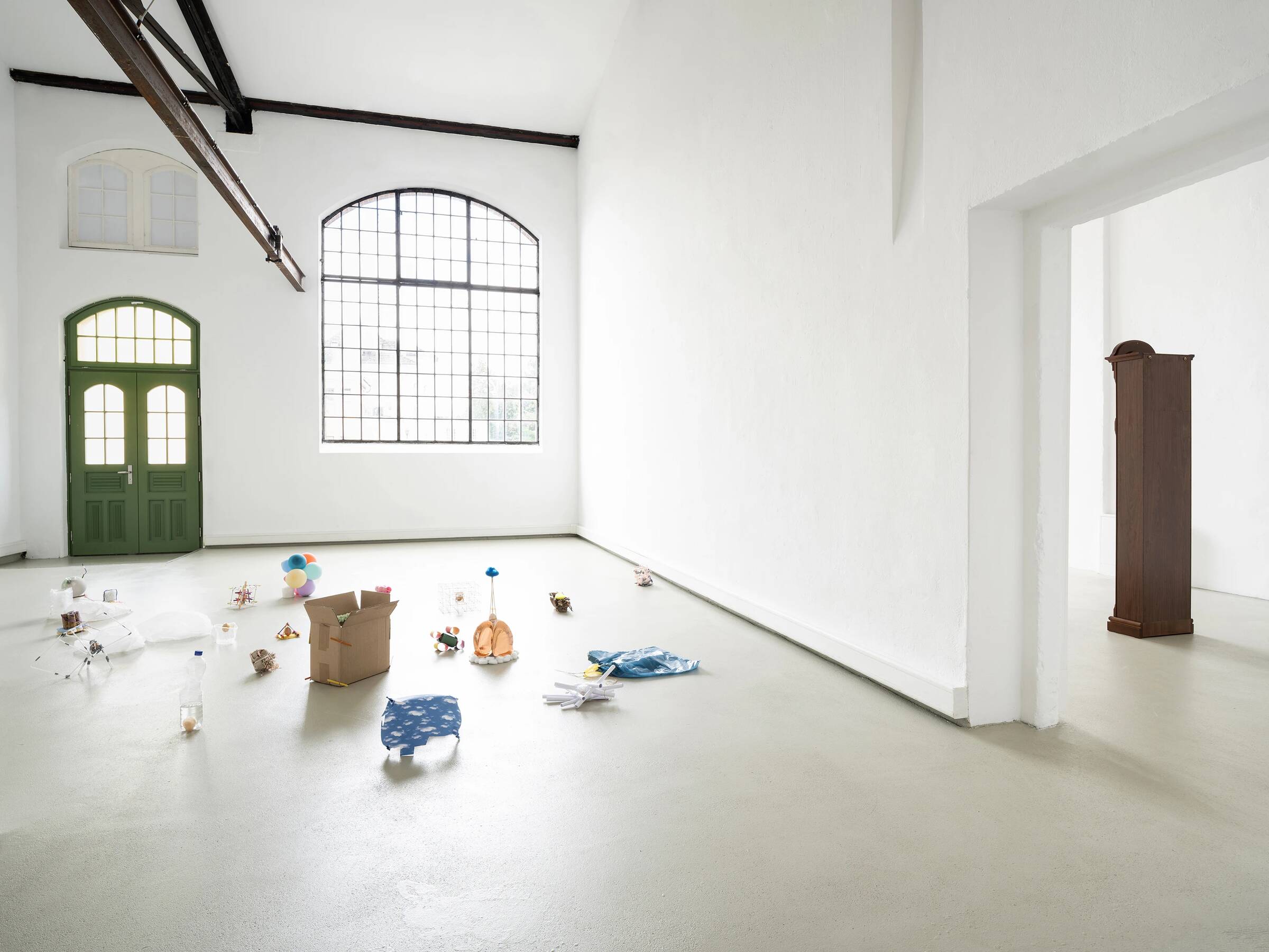 Helle Ausstellungsansicht mit weiß gestrichenen Wänden und Boden. Im Vordergrund stehen verstreut verschiedene kleine Objekte und Skulpturen, darunter ein Karton, ein blauer Gegenstand mit Muster, eine orangefarbene Form und weitere bunte Elemente. Links befindet sich eine grün gestrichene Doppeltür mit Oberlicht, darüber ein großes, rechteckiges Fenster mit Rundbogen. Rechts ist ein Durchgang zu einem weiteren Raum sichtbar, in dem eine hohe, schmale, braune Standuhr steht.
