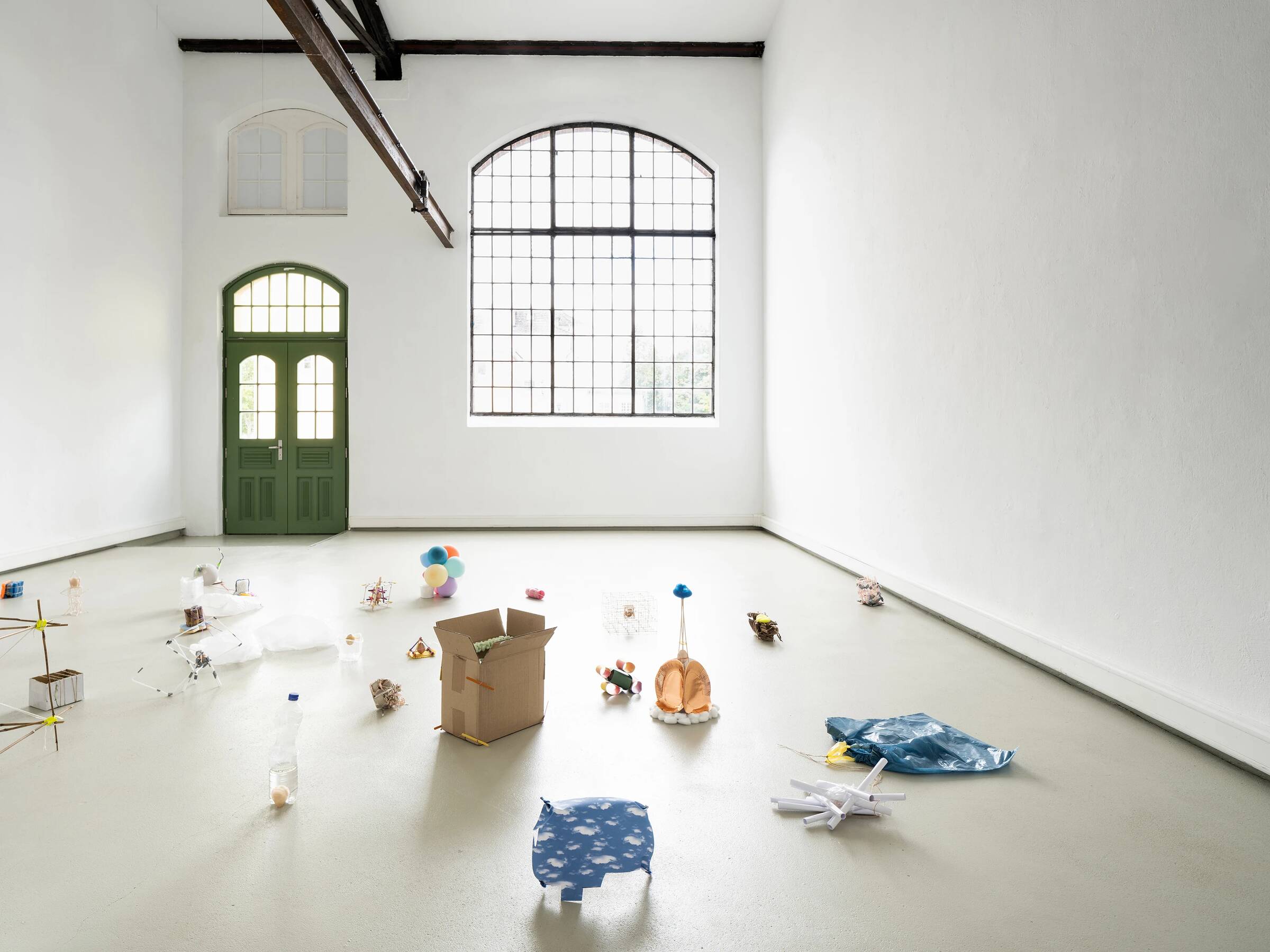 Weißer Ausstellungsraum mit hohen Wänden und großen Fenstern. Im Raum verteilt stehen verschiedene kleine Objekte und Skulpturen auf dem Boden, darunter ein brauner Karton, eine blaue Plastikplane, eine durchsichtige Plastikflasche, ein kleines Modell mit bunten Luftballons und weitere abstrakte Formen. Die Tür ist grün und hat ein Fenster mit Rundbogen. Die Decke zeigt sichtbare Holzbalken.