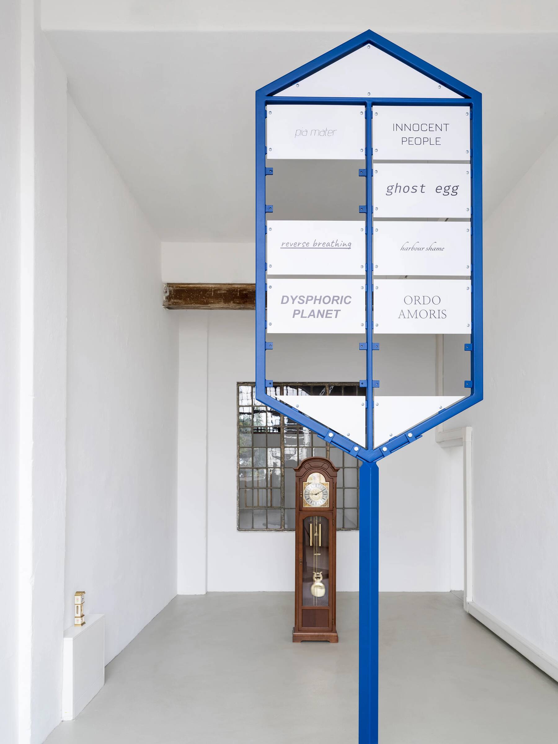 Eine blaue, pfeilförmige Standskulptur mit mehreren weißen Tafeln, die verschiedene Texte tragen: 'pia mater', 'INNOCENT PEOPLE', 'ghost egg', 'reverse breathing', 'harbour shame', 'DYSPHORIC PLANET' und 'ORDO AMORIS'. Im Hintergrund steht eine alte Standuhr vor einer weißen Wand mit einem Fenster.