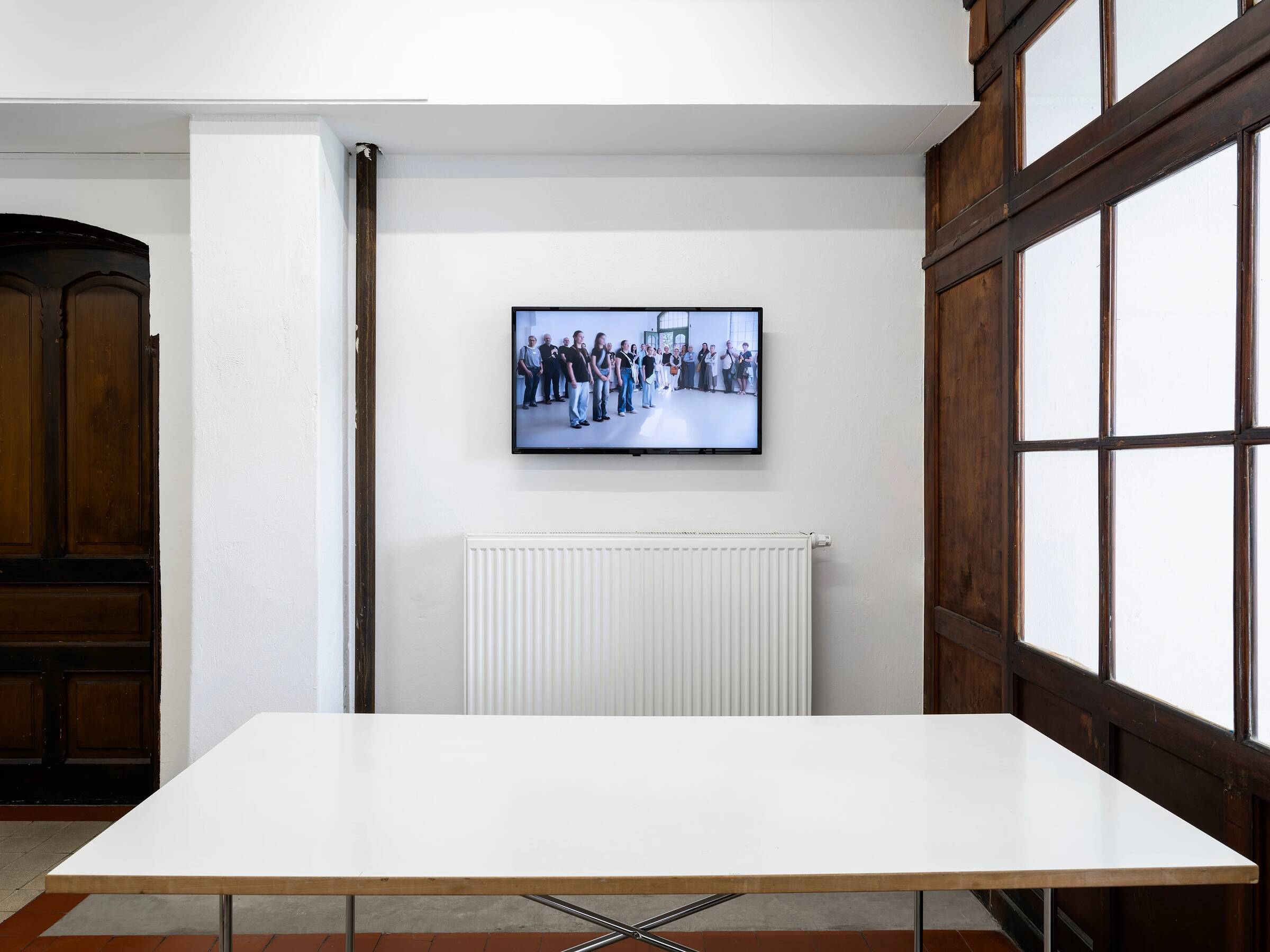 Weißer Tisch im Vordergrund, dahinter weiße Wand mit Heizkörper und einem Bildschirm, der eine Gruppe von Menschen zeigt.