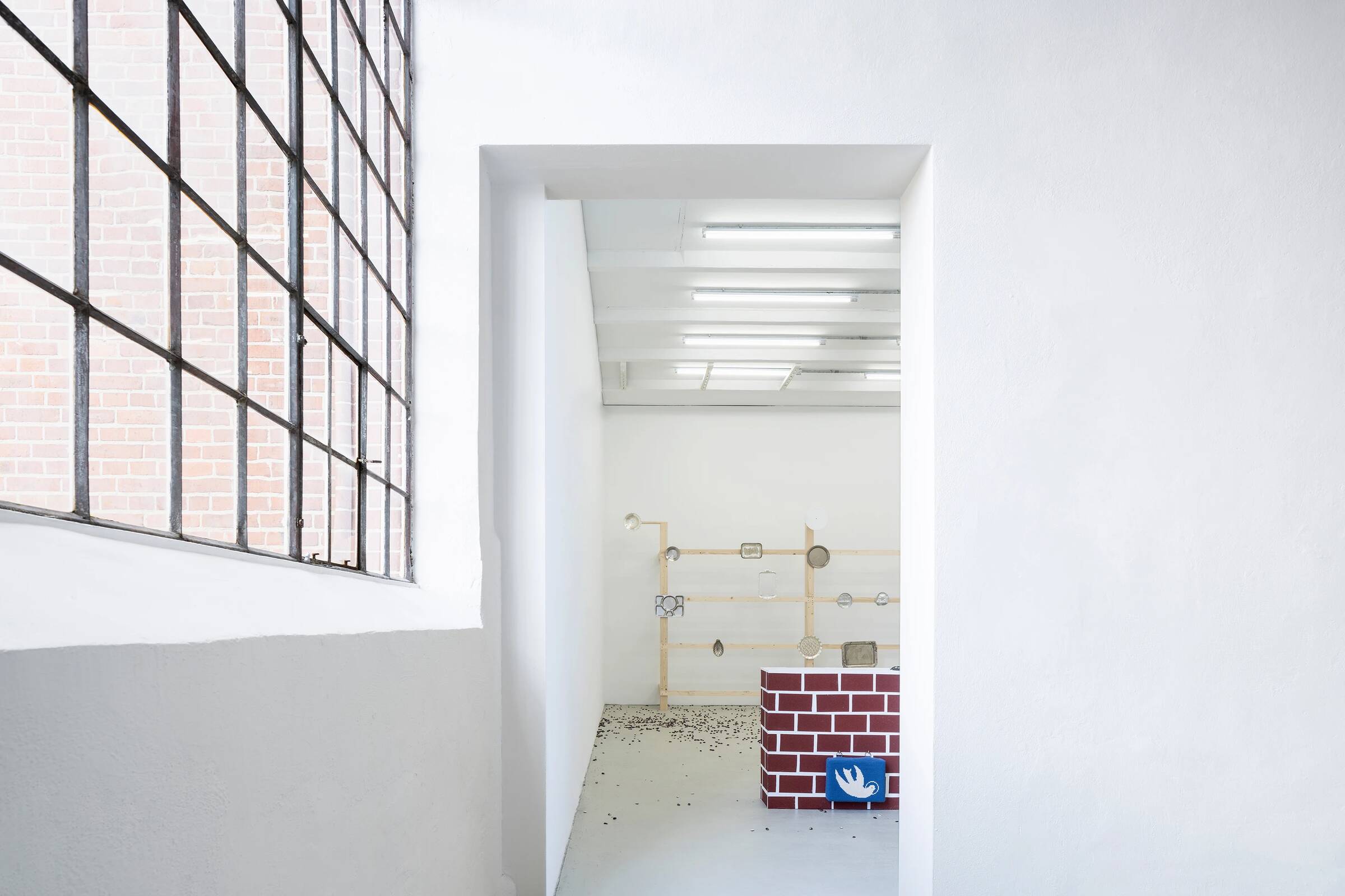 Blick durch einen weißen Türrahmen in einen Ausstellungsraum. Im Vordergrund rechts steht ein niedriger, rechteckiger Kasten mit einer rot-weißen Ziegelsteinmusterung. Darauf liegt ein gestricktes, blaues Schild mit einer weißen Friedenstaube. Im Hintergrund ist eine Wand mit einer minimalistischen Installation aus Holzstäben und runden Elementen zu sehen. Der Boden ist weiß, und die Decke hat mehrere lange, schmale Leuchtstofflampen. Links ist ein großes Fenster mit einem schwarzen Metallgitter und dahinter eine rote Backsteinwand sichtbar.