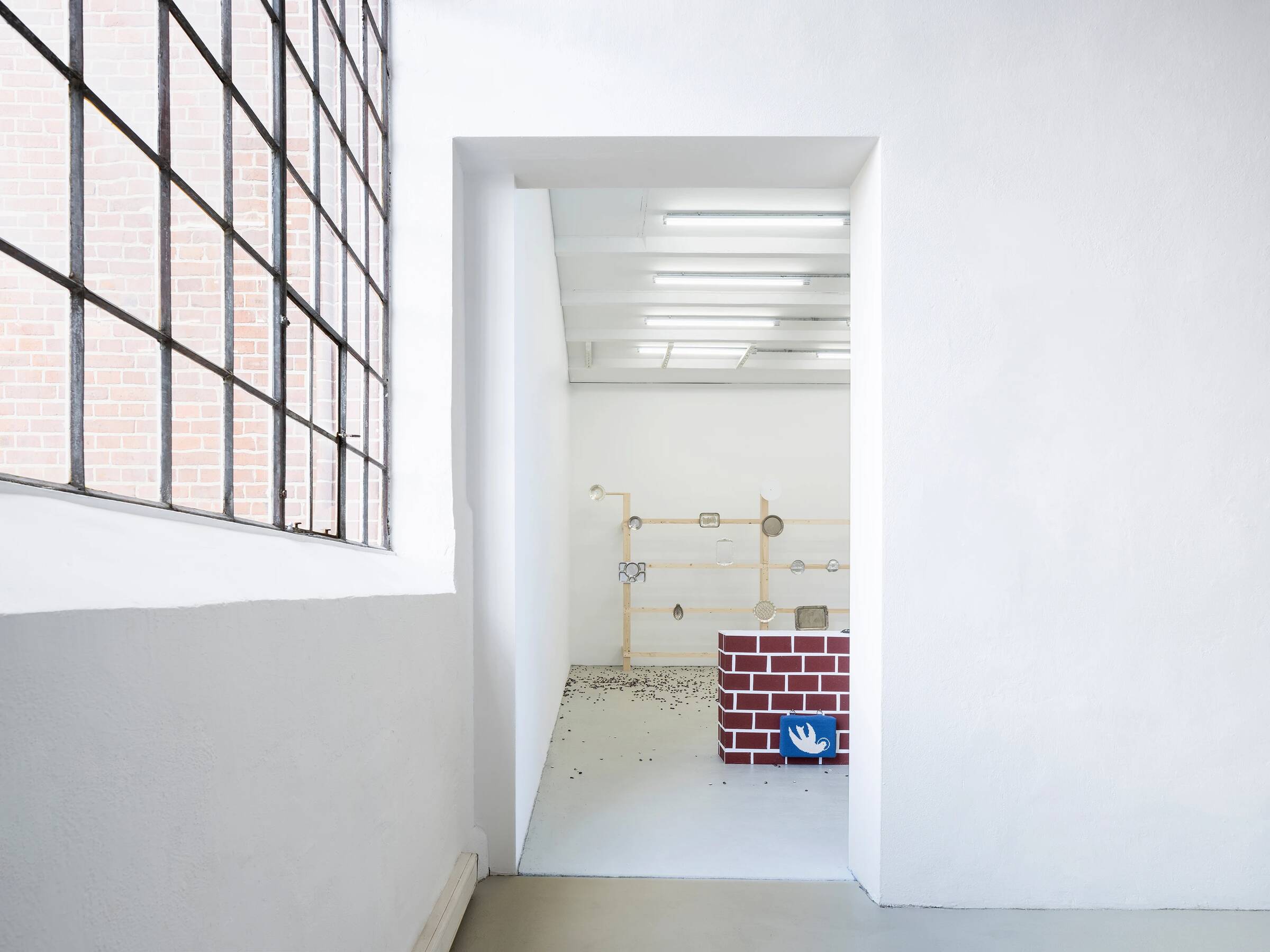 Blick durch einen weißen Türrahmen in einen Ausstellungsraum. Im Vordergrund rechts steht ein niedriger, rechteckiger Kasten mit einer rot-weißen Ziegelsteinmusterung. Darauf liegt ein gestricktes, blaues Schild mit einer weißen Friedenstaube. Im Hintergrund ist eine Wand mit einer minimalistischen Installation aus Holzstäben und runden Elementen zu sehen. Der Boden ist weiß, und die Decke hat mehrere lange, schmale Leuchtstofflampen. Links ist ein großes Fenster mit einem schwarzen Metallgitter und dahinter eine rote Backsteinwand sichtbar.