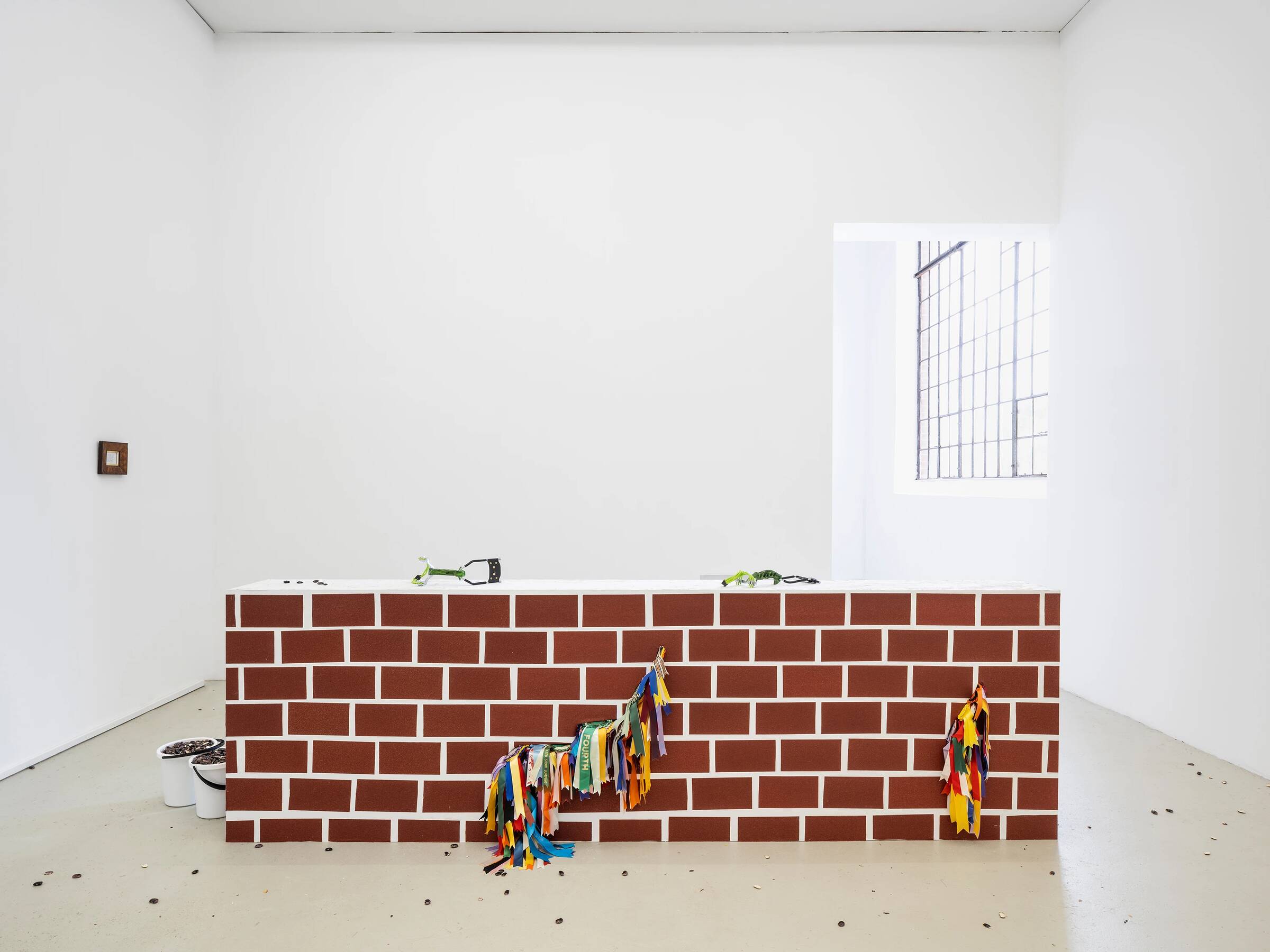 Raum mit weißer Wand und Fenster, davor ein quaderförmiges Objekt mit rot-weißer Ziegelsteinoptik, an dem bunte Stoffstreifen hängen.