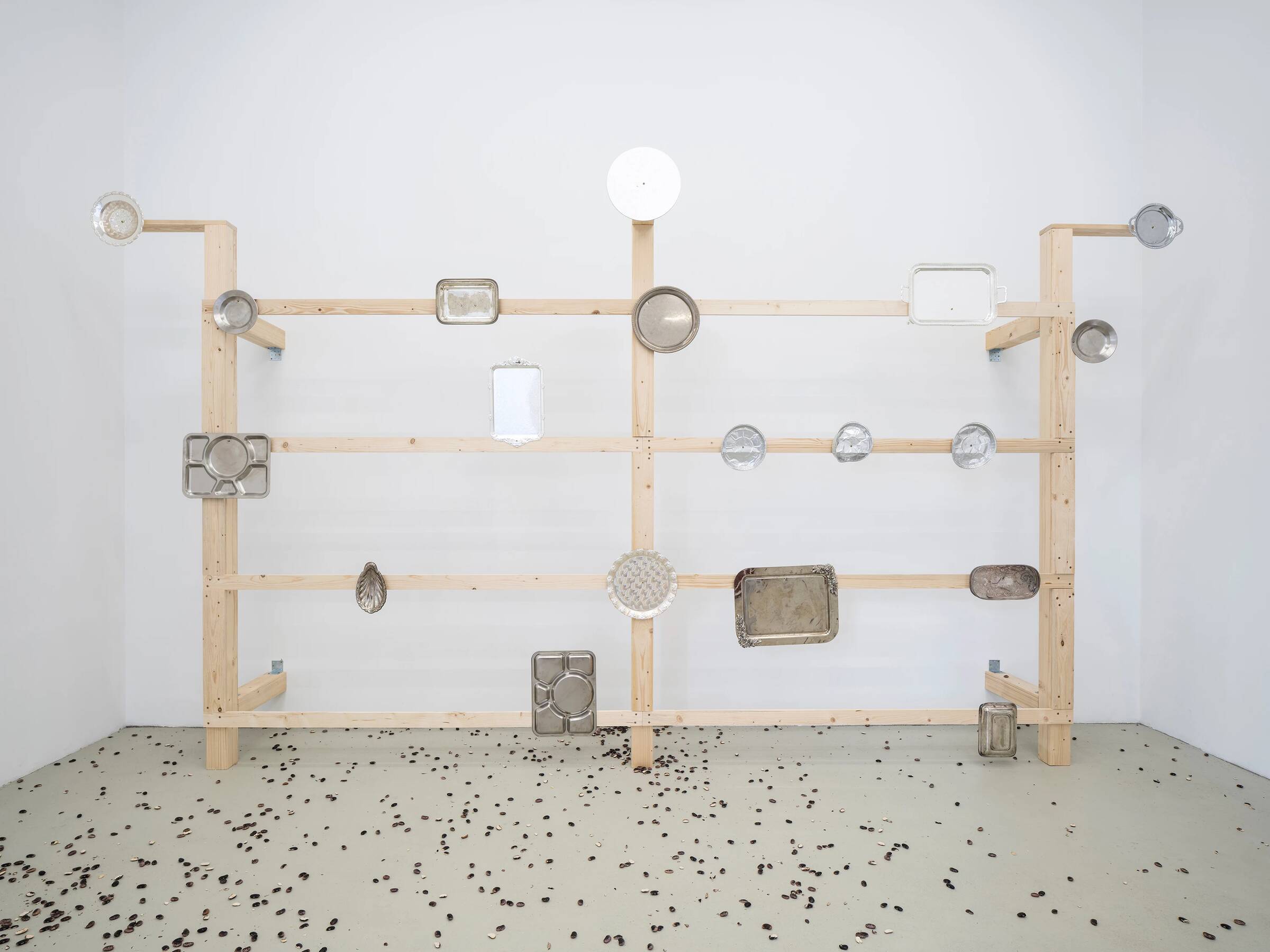 Holzgestell mit mehreren waagerechten und senkrechten Latten, an denen verschiedene Metallbackformen in unterschiedlichen Formen und Größen hängen, auf weißem Boden mit verstreuten kleinen schwarzen Objekten.