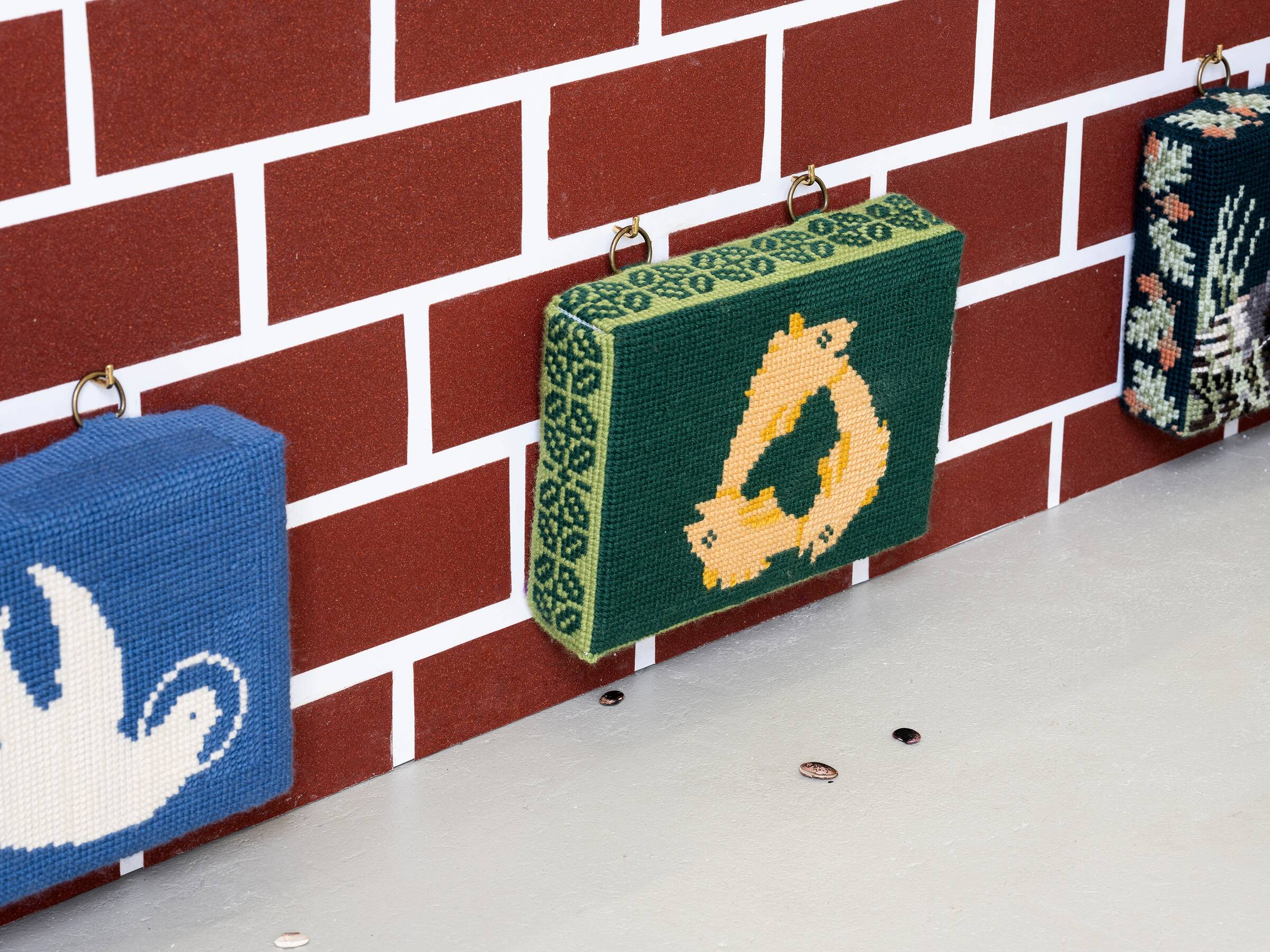 Drei quadratische, gehäkelte Wandbehänge mit Tiermotiven hängen an einer rot-weißen Ziegelsteinwand, darunter liegt ein weißer Boden mit kleinen Bohnen.
