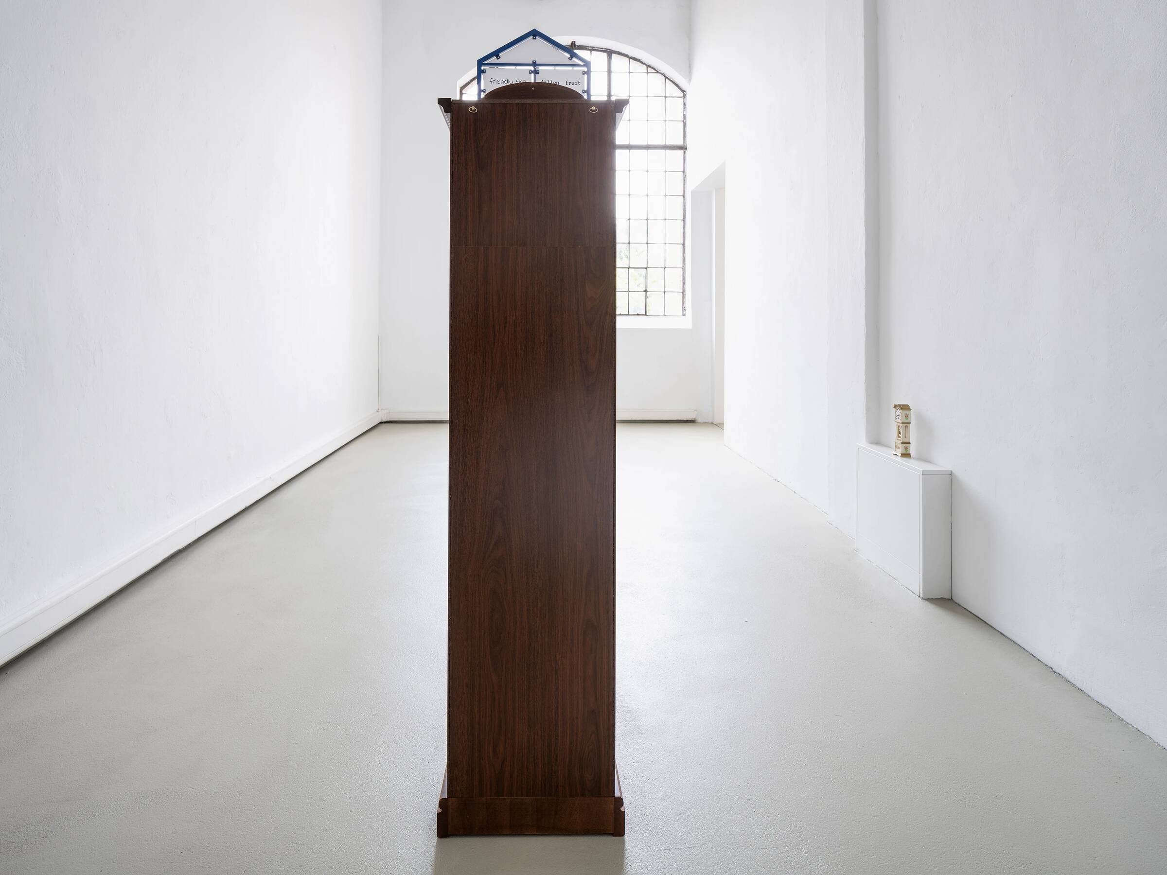 In einem weißen, hellen Ausstellungsraum steht mittig eine hohe, schmale Standuhr aus dunklem Holz. Im Hintergrund ist ein großes, rechteckiges Fenster mit einem Gittermuster zu sehen. Rechts an der Wand steht ein kleiner weißer Sockel mit einem kleinen Modell einer Standuhr darauf.