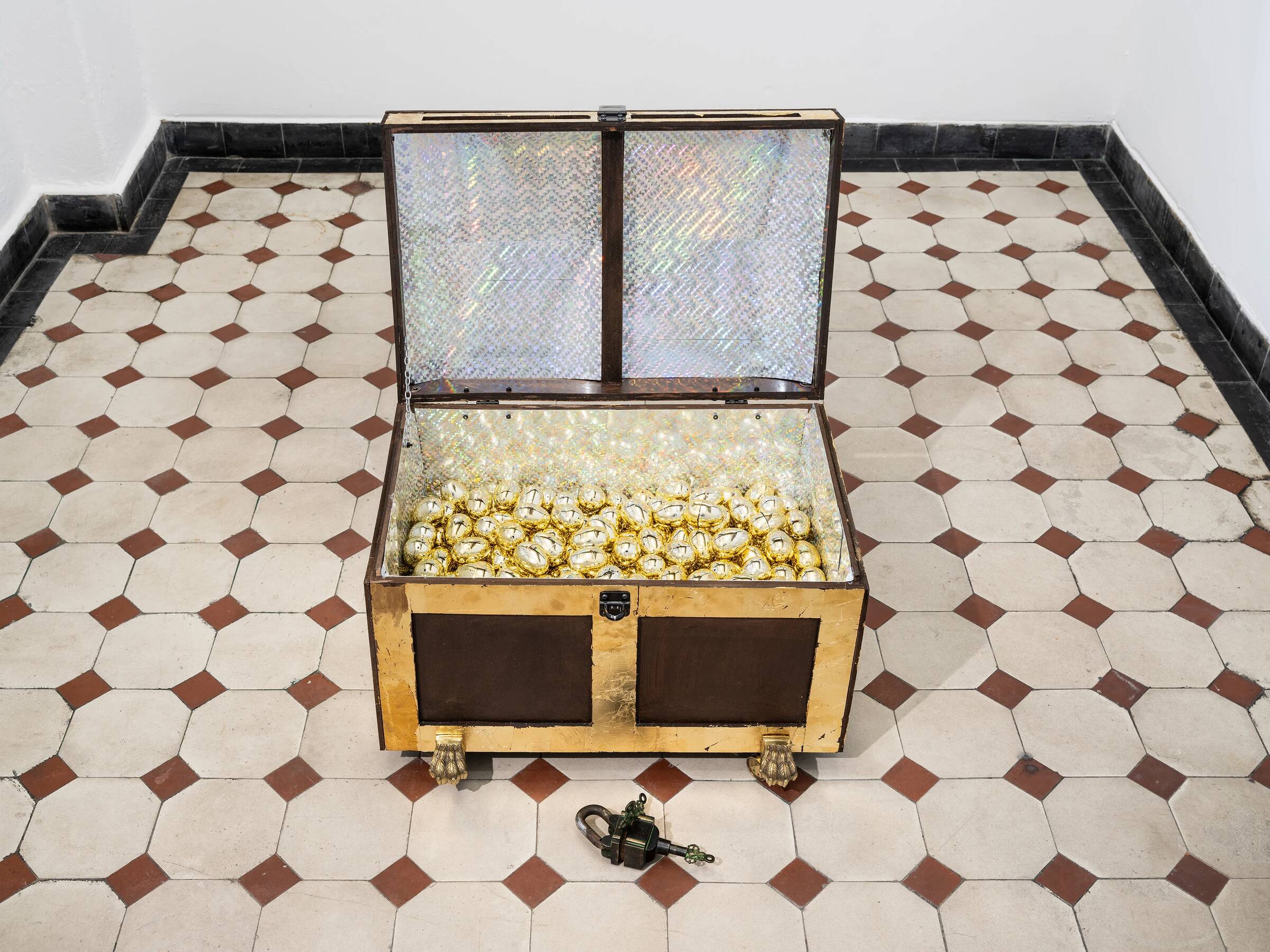 Ein offener, rechteckiger Schatzkoffer mit goldfarbenen und braunen Flächen steht auf einem weiß-braun gemusterten Boden. Im Inneren des Koffers liegen viele goldene Kugeln. Vor dem Koffer liegt ein großes, antik wirkendes Vorhängeschloss.