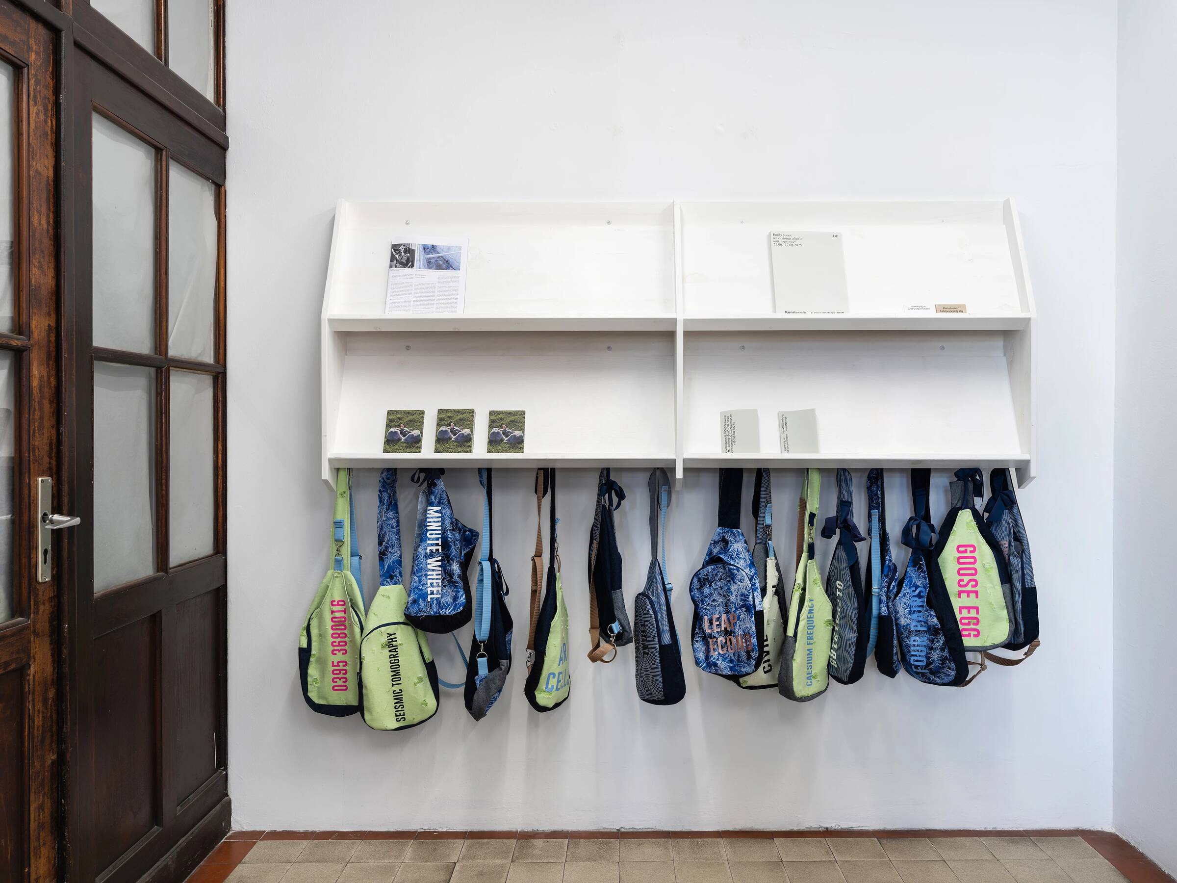 Wandinstallation mit mehreren blauen und grünen Taschen, teils mit Aufdrucken wie 'GOOSE EGG', unter zwei weißen Regalen mit kleinen Texttafeln, neben einer Holztür.