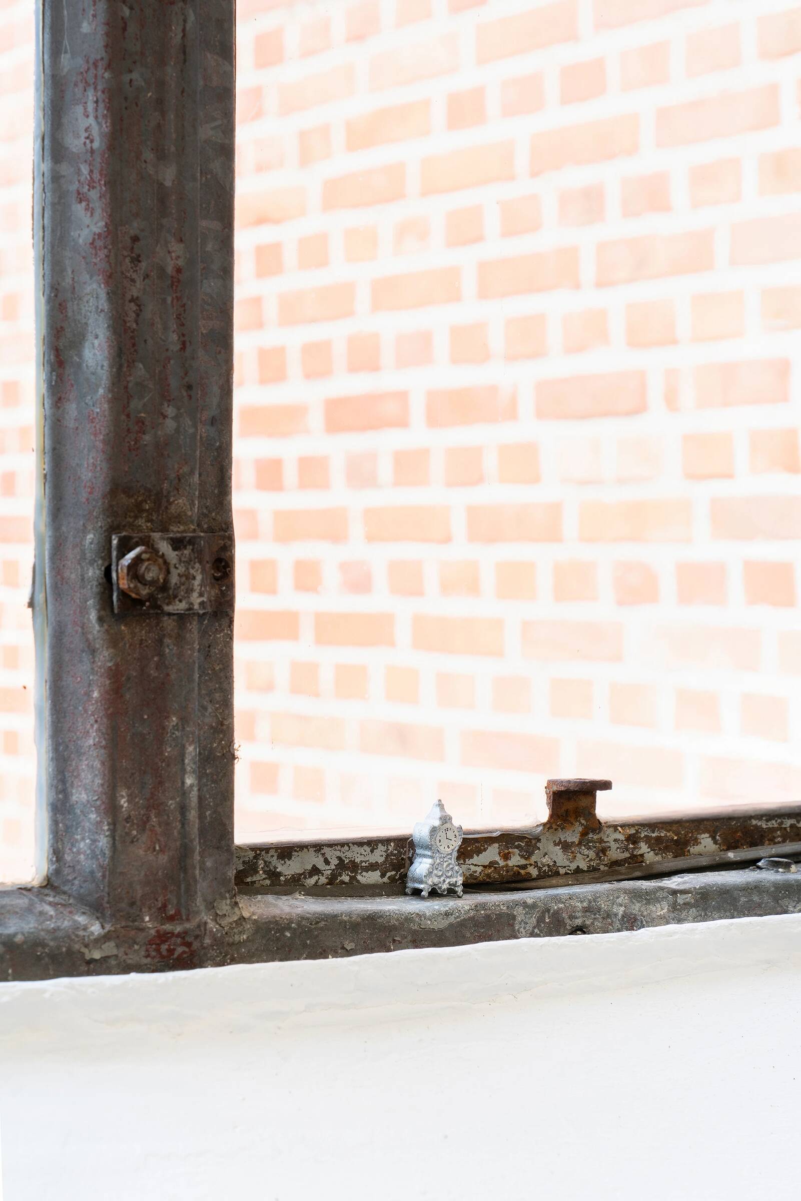 Eine sehr kleine, silberfarbene, detailliert gearbeitete Standuhr steht auf einer weißen Fensterbank. Dahinter ist ein rostiger, metallener Fensterrahmen mit einer Schraube sichtbar. Im Hintergrund ist eine unscharfe, hellrote Backsteinmauer zu sehen.